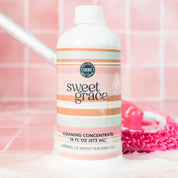 A bottle of Bridgewater Candle Sweet Grace Cleaning Concentrate | Mop Soap rests on a pink tiled surface with cleaning foam, while a pink mop in the background showcases this fragrant cleaner’s stylish appeal.