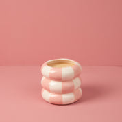 The Bridgewater Candle Sweet Grace Collection Candle #074, featuring a round, pink and white striped, doughnut-like design, sits on a pink surface with a matching pink background.