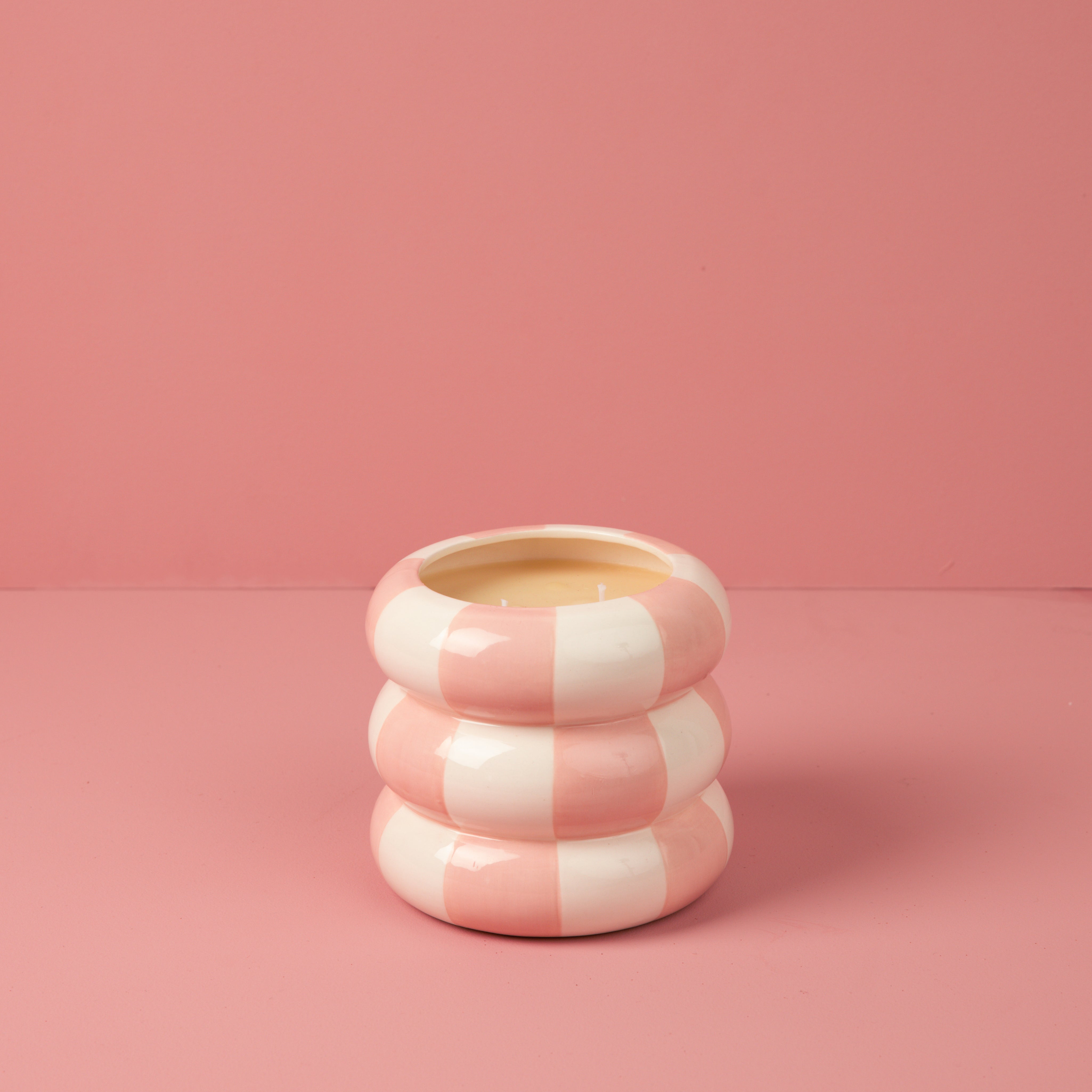 The Bridgewater Candle Sweet Grace Collection Candle #074, featuring a round, pink and white striped, doughnut-like design, sits on a pink surface with a matching pink background.