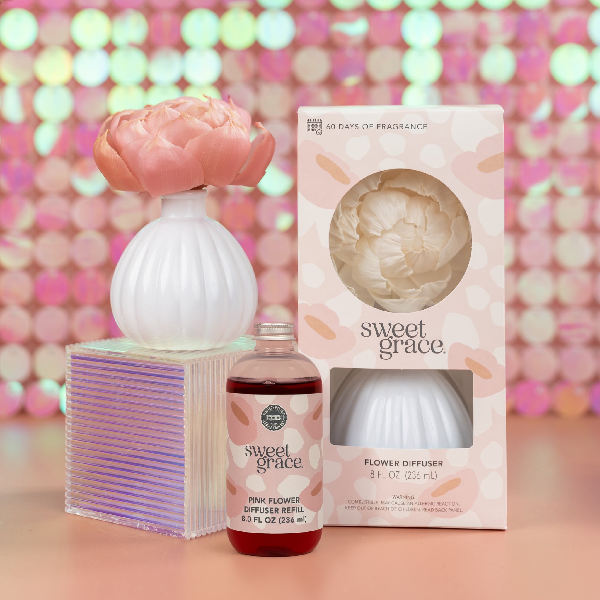 The Bridgewater Candle Sweet Grace Pink Dye Fragrance Refill Oil is displayed with its product box and a white flower-shaped diffuser topped with a pink flower, all set against a pink bokeh background for an elegant presentation.