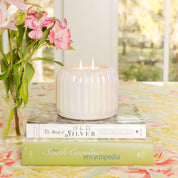 A white Bridgewater Candle Sweet Grace Collection Candle #071 with two flames sits atop stacked books beside a vase of pink flowers on a floral tablecloth, filling the air with fragrance, with a window and greenery in the background.