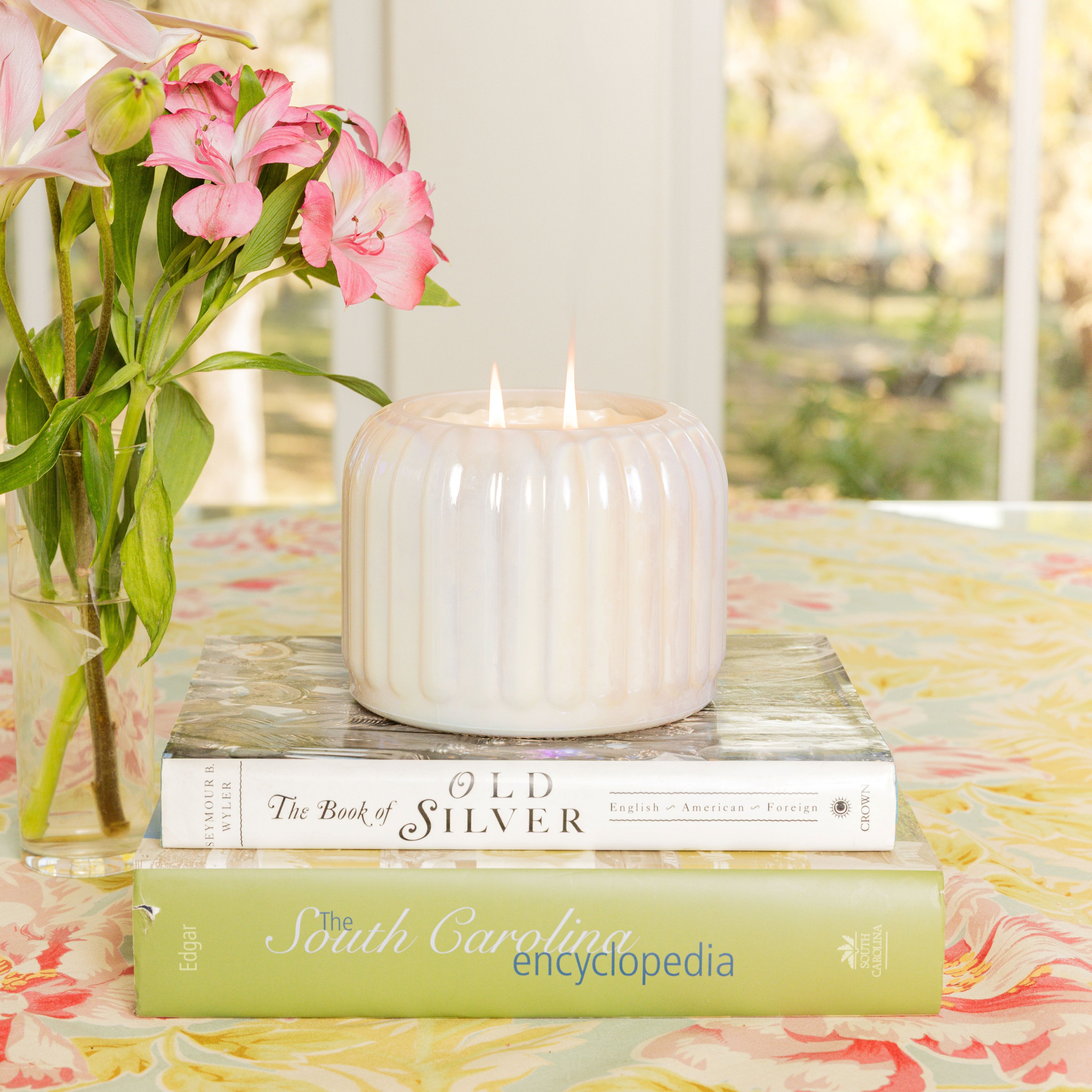 A white Bridgewater Candle Sweet Grace Collection Candle #071 with two flames sits atop stacked books beside a vase of pink flowers on a floral tablecloth, filling the air with fragrance, with a window and greenery in the background.