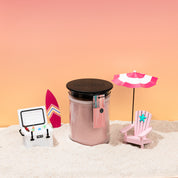 The Bridgewater Candle Sunkissed 18oz Large Jar Candle sits on sand, surrounded by a toy cooler with drinks, a small surfboard, a mini pink beach chair, and a pink-and-white umbrella—all set against a pink-orange gradient backdrop.