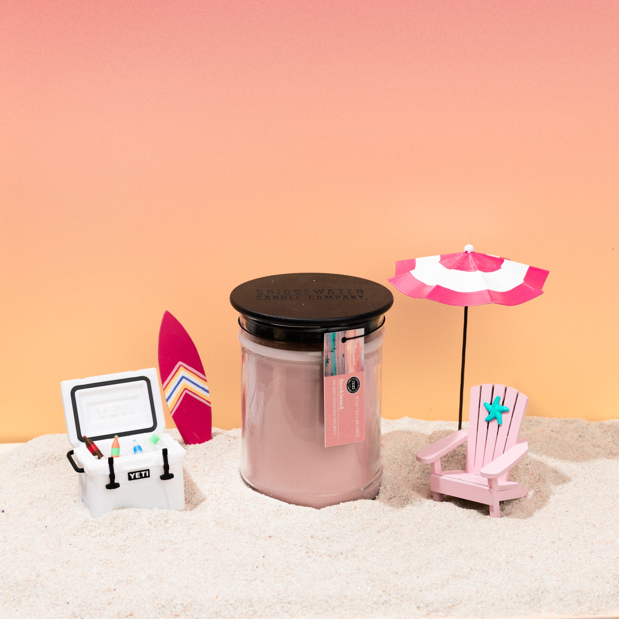 The Bridgewater Candle Sunkissed 18oz Large Jar Candle sits on sand, surrounded by a toy cooler with drinks, a small surfboard, a mini pink beach chair, and a pink-and-white umbrella—all set against a pink-orange gradient backdrop.