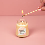 A hand lights the Bridgewater Candle Sweet Grace Collection Candle #078, crafted with soy-blend wax in a petite glass jar with a pastel label, set against a pink background.