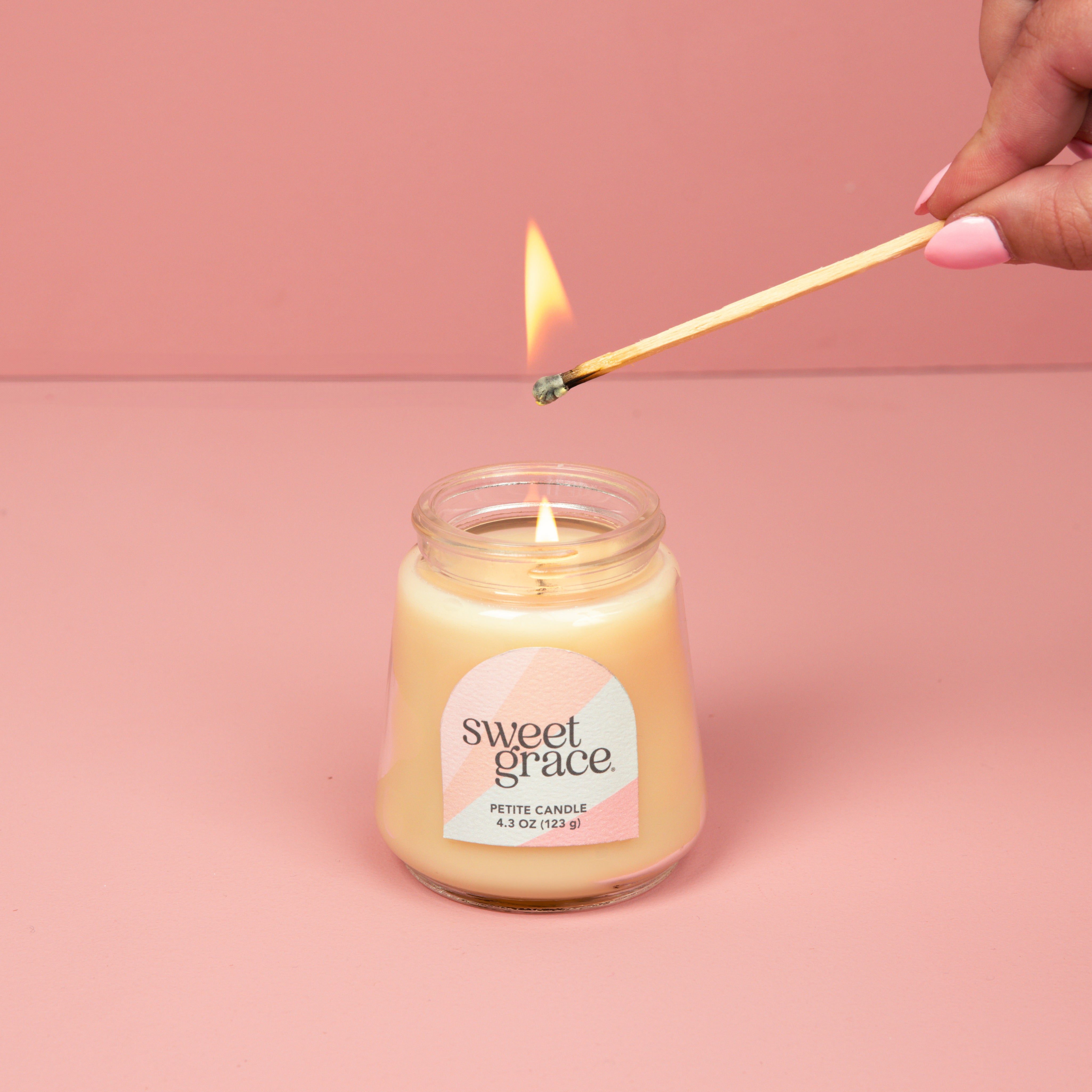 A hand lights the Bridgewater Candle Sweet Grace Collection Candle #078, crafted with soy-blend wax in a petite glass jar with a pastel label, set against a pink background.