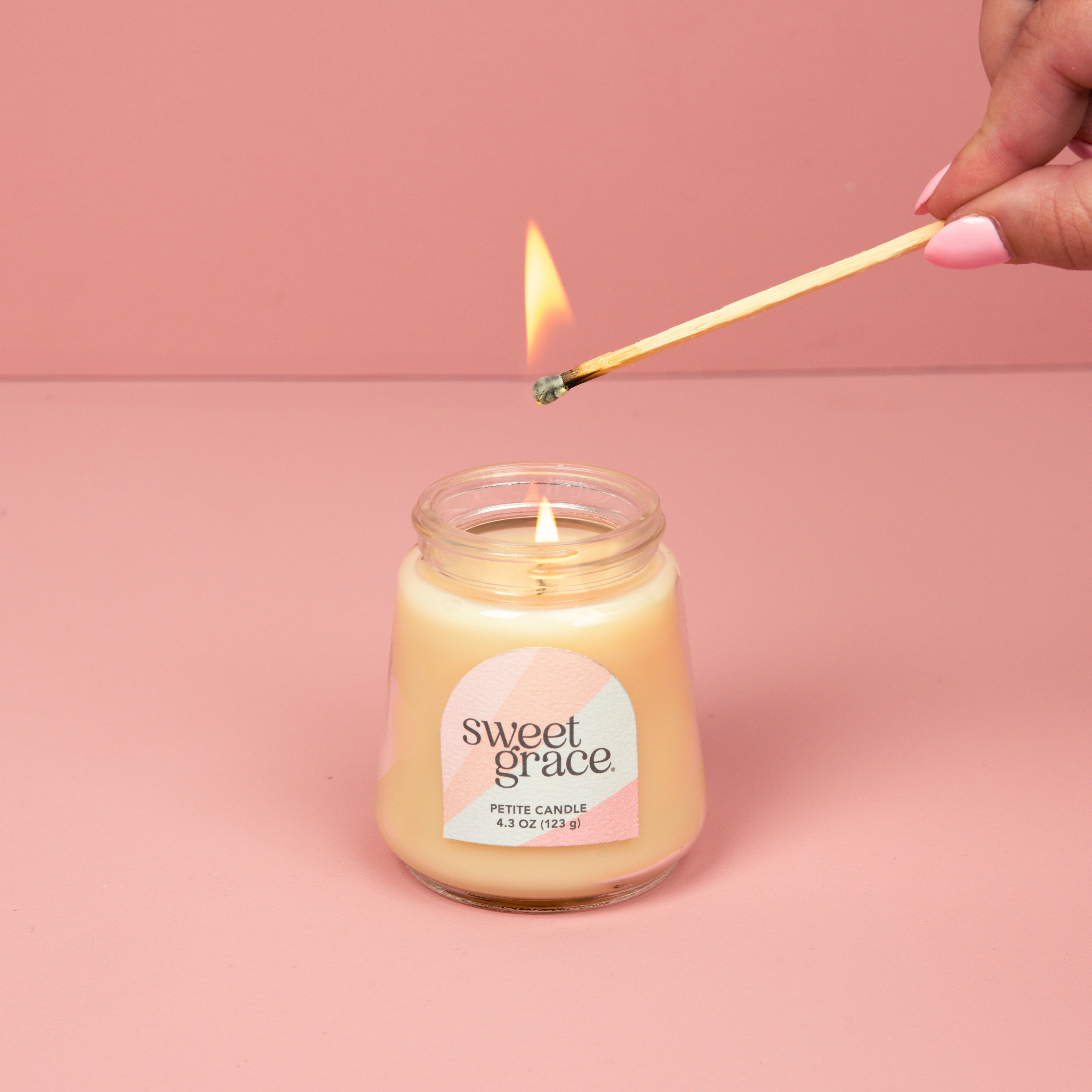 A hand lights the Bridgewater Candle Sweet Grace Collection Candle #078, crafted with soy-blend wax in a petite glass jar with a pastel label, set against a pink background.