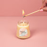 A hand lights the Bridgewater Candle Sweet Grace Collection Candle #078, crafted with soy-blend wax in a petite glass jar with a pastel label, set against a pink background.