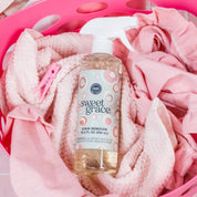 A bottle of Bridgewater Candle's Sweet Grace Stain Remover sits on pink towels in a pink laundry basket, with a pink clothespin nearby, showcasing this gentle fabric and upholstery cleaner in a warm, colorful setting.