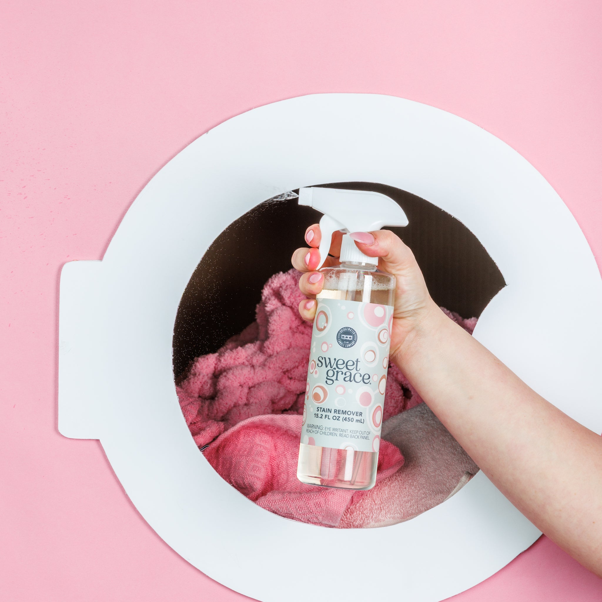 A hand sprays Bridgewater Candle's Sweet Grace Stain Remover onto pink towels inside a white washing machine door, set against a pink background.