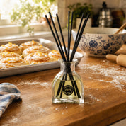 The Bridgewater Candle Rise & Shine Reed Diffuser with black reeds sits on a floured wooden kitchen counter, filling the air with a cozy cinnamon roll scent as pastries and sunlight complete the inviting scene.