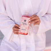 A person wearing a white bathrobe holds a clear bottle of Bridgewater Candle Sweet Grace Bubble Bath featuring a label with white and pink circles.