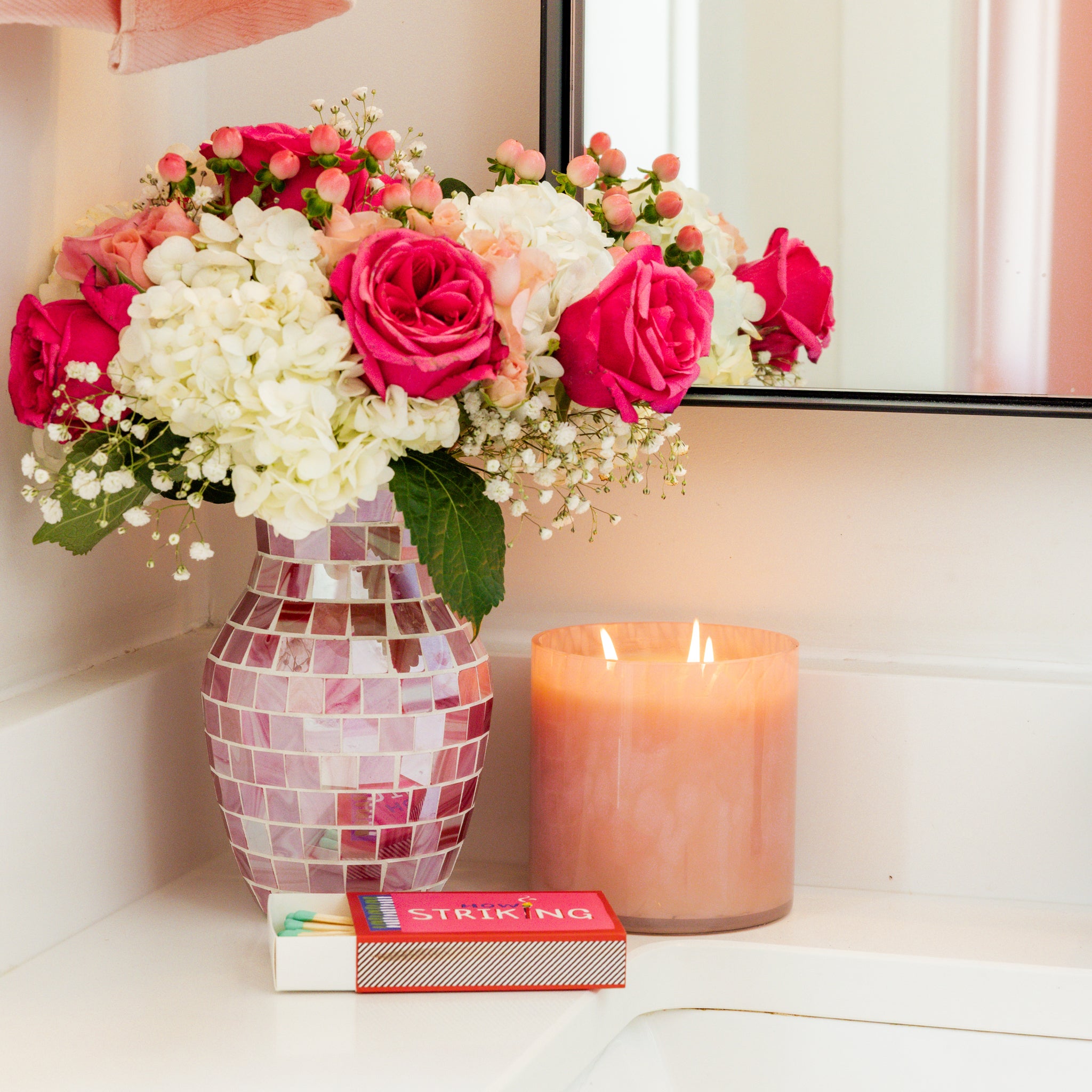 A pink mosaic vase with white and pink flowers sits on a bathroom counter beside a lit Bridgewater Candle Sweet Grace Collection Candle #073, a box of matches, and a mirror reflecting part of the arrangement.