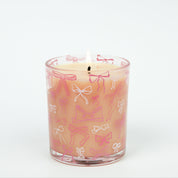 The Sweet Grace Pink Bow Candle by Bridgewater Candle features delicate white and pink bow designs on a glass holder. Made with soy-blend wax, this charming candle is displayed against a plain white background.