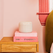 A white ribbed Bridgewater Candle Sweet Grace Collection Candle #071 sits on two pink books atop a light wood bedside table, with a pale pink wall, red window frame, and green upholstered headboard in the background.