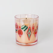 A lit Sweet Grace Ornament Decal Candle by Bridgewater Candle, featuring light cream soy-blend wax in a clear glass holder adorned with colorful, retro-style ornament designs against a white background.