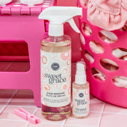 Two bottles of Bridgewater Candle’s Sweet Grace Stain Remover rest on pink tiles next to a pink laundry basket filled with clothes and a pink clothespin.
