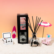 A beach scene displays the Bridgewater Candle Sunkissed Reed Diffuser with fiber reeds, surrounded by a cooler, surfboard, pink and white umbrella, and a pink chair on sand against a pink-orange gradient backdrop.