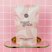 A 2 lb (907 g) pink and white bag of Bridgewater Candle Sweet Grace Soaking Salts, made with Pink Himalayan sea salt, sits on a glass shelf against a pink tiled wall with gold brackets.