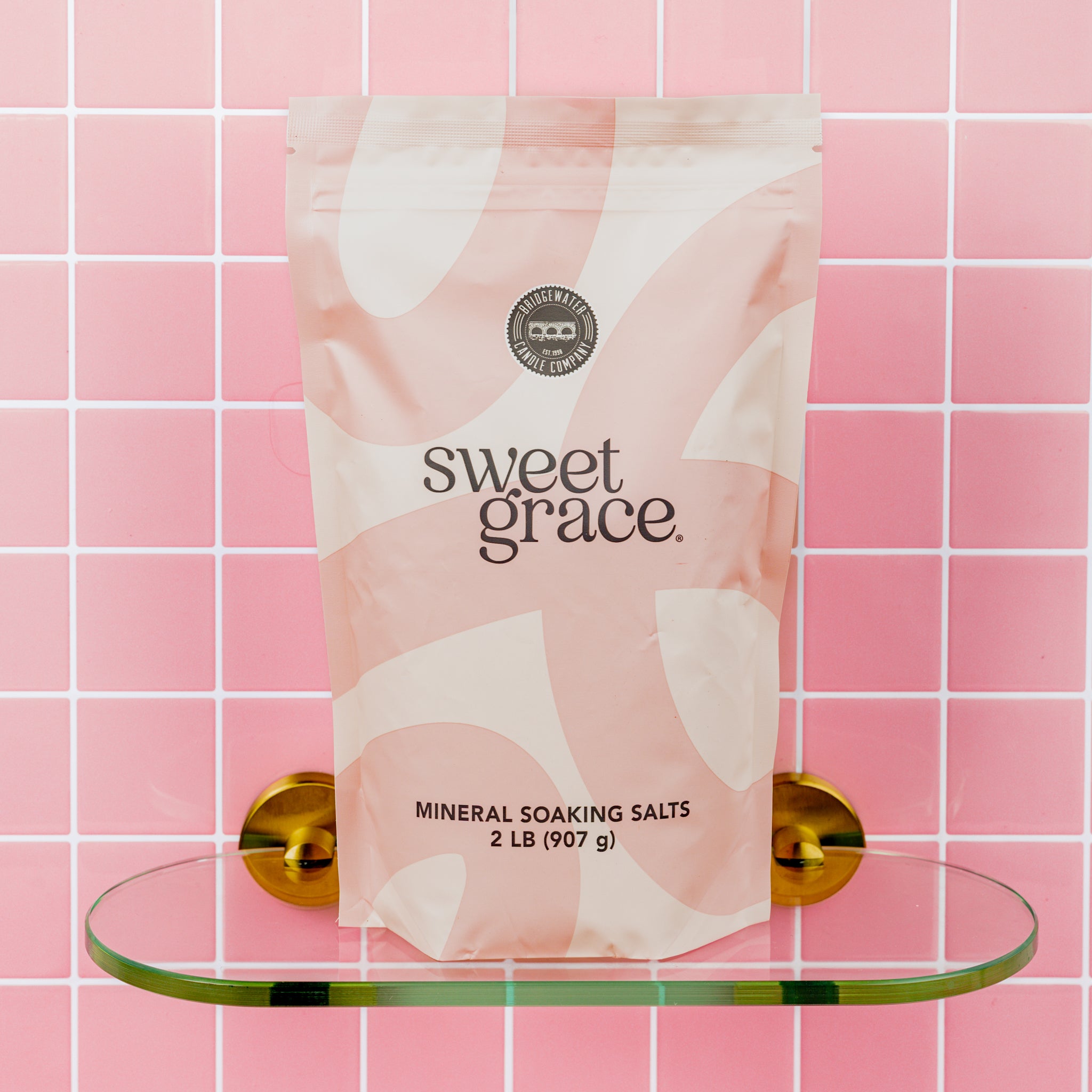 A 2 lb (907 g) pink and white bag of Bridgewater Candle Sweet Grace Soaking Salts, made with Pink Himalayan sea salt, sits on a glass shelf against a pink tiled wall with gold brackets.