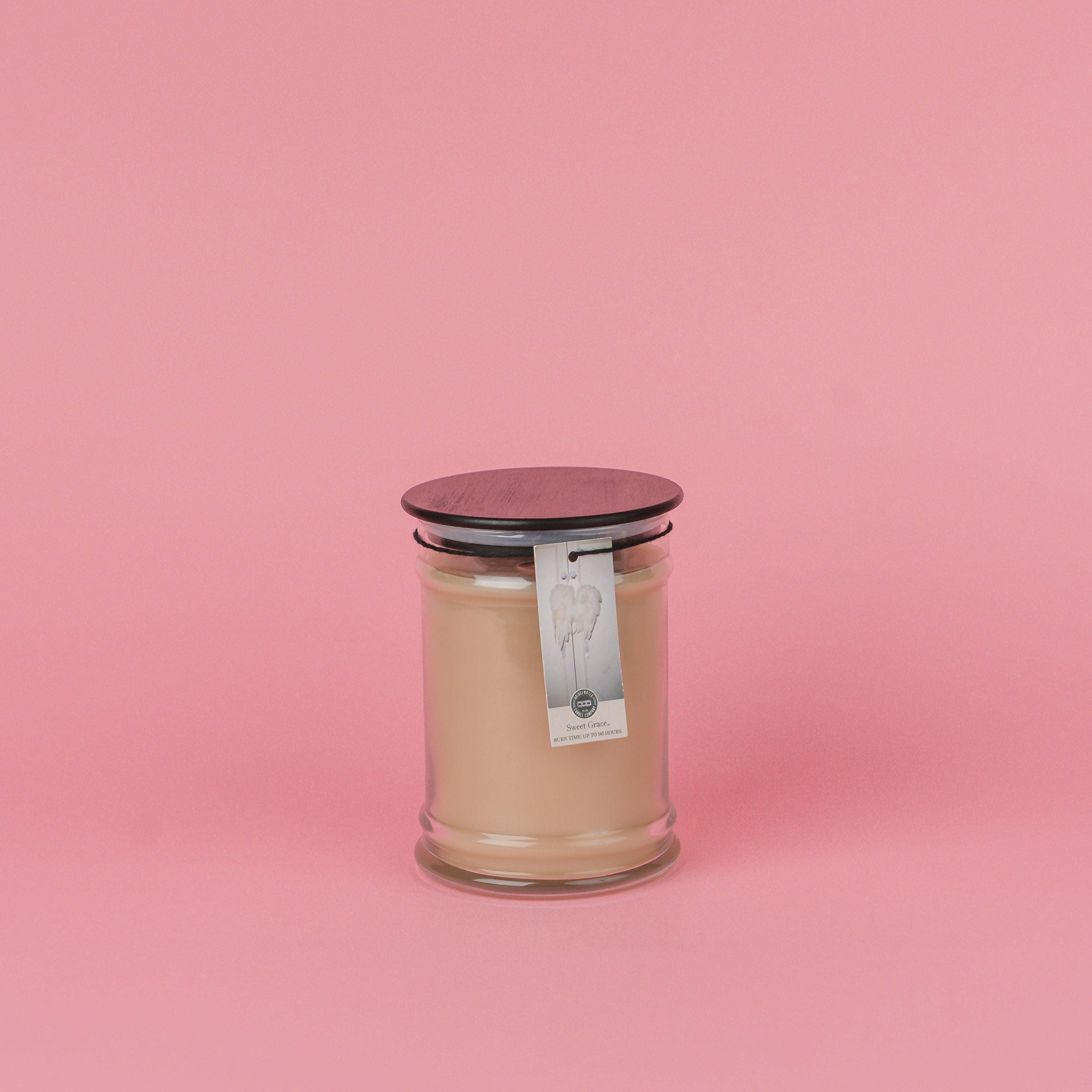 A beige Bridgewater Candle Sweet Grace 18oz Large Jar Candle with a dark lid sits on a pink background. An angel wings label hangs from the lid, highlighting the signature Sweet Grace fragrance.