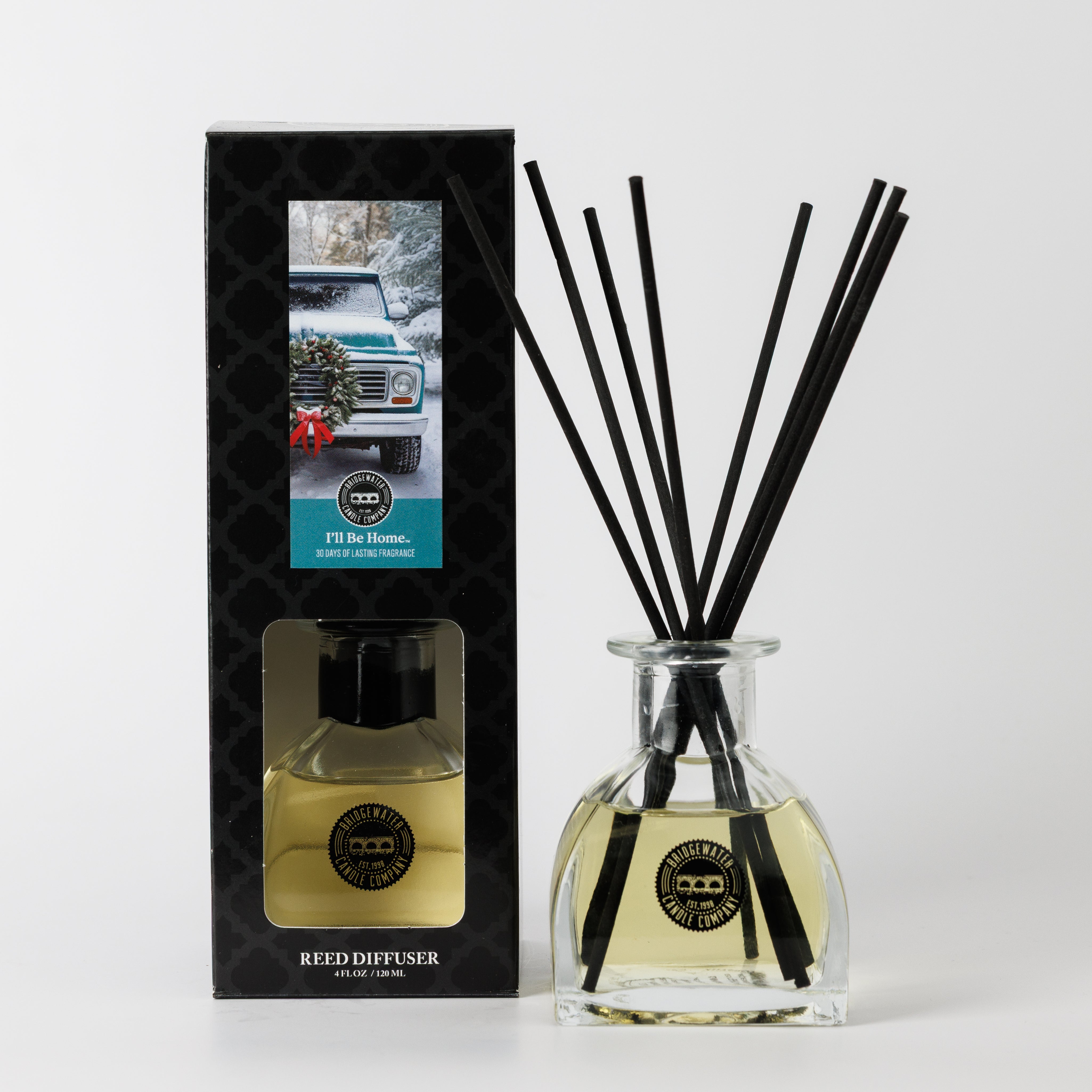 The Bridgewater Candle "I'll Be Home" Reed Diffuser features sleek black reeds in a glass bottle beside packaging with a wreath-adorned car in snow, promising a welcoming home fragrance with its pale yellow liquid.