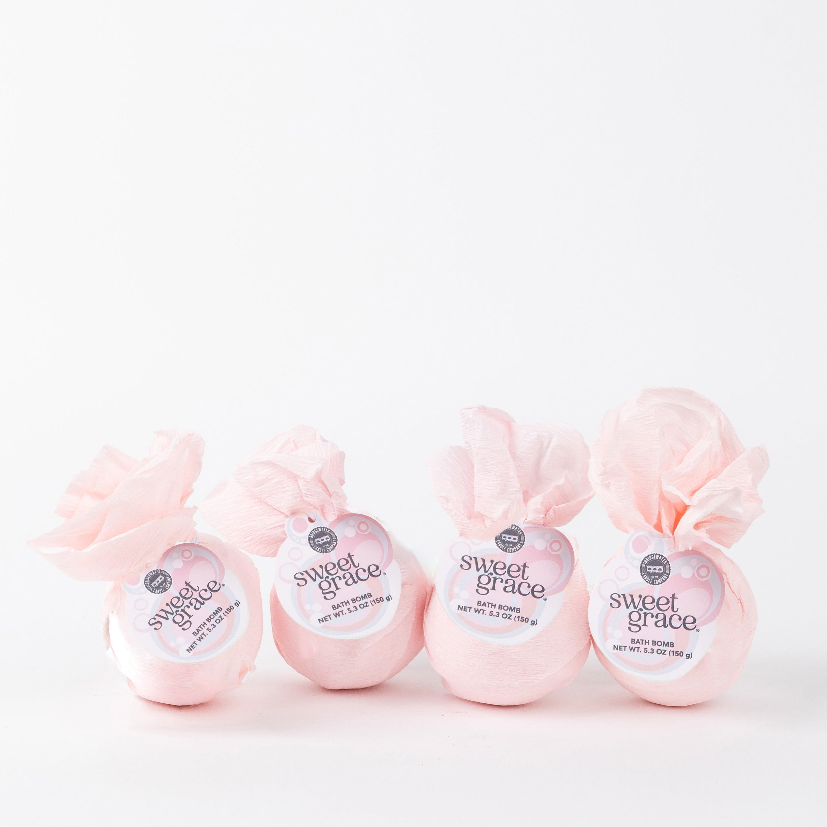 Four Bath Bomb 4 Pack-Sweet Grace by Bridgewater Candle, wrapped in light pink paper and featuring the best-selling Sweet Grace scent, are lined up against a plain white background.
