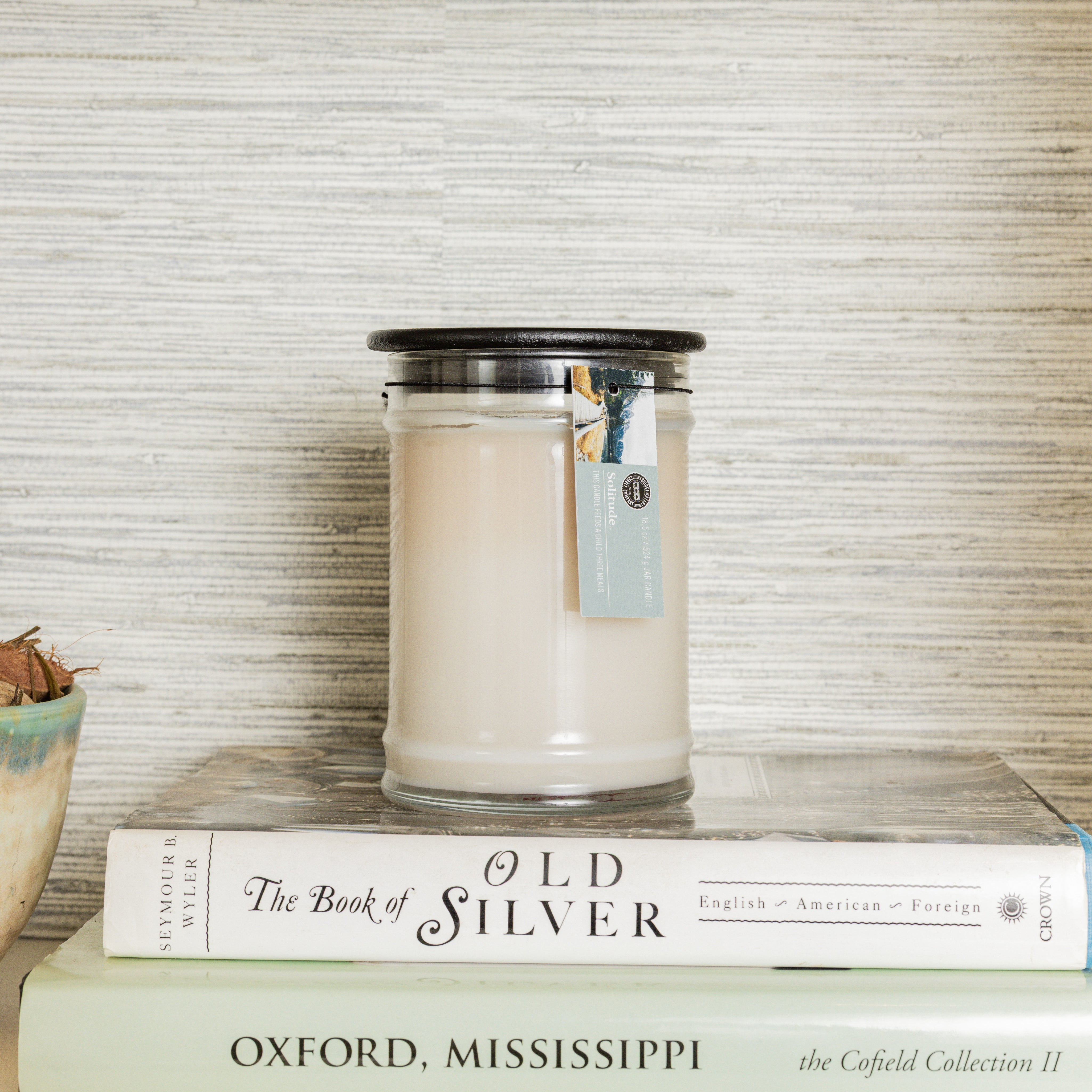 The Solitude Large Jar Candle by Bridgewater Candle, featuring soy-blend wax in a glass jar with a black lid and tag, sits atop two stacked books—The Book of Old Silver and Oxford, Mississippi—beside a small ceramic bowl with a plant.