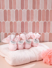 Bridgewater Candles Bath Bomb 4 Pack-Sweet Grace, the brand’s best-selling scent, comes wrapped in tissue with labels and displayed on a light pink towel beside a matching bath pouf, creating a luxurious spa-inspired setting against pink tiles.