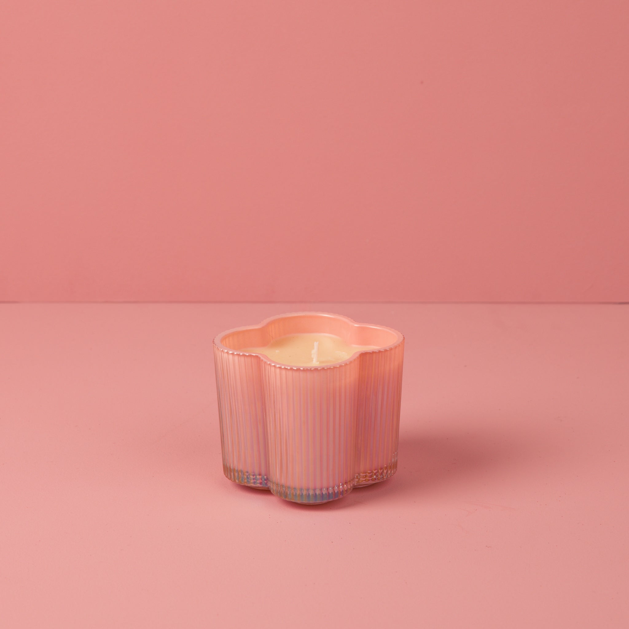 A pastel pink, flower-shaped vessel holds Bridgewater Candle's Sweet Grace Collection Candle #075, its soy-blend wax glows softly on a light pink surface against a matching background.