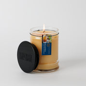A lit Rise & Shine 8oz Jar Candle by Bridgewater Candle, with a black lid and blue tag, contains light beige soy-blend wax and signature fragrance. The candle sits against a plain white background.