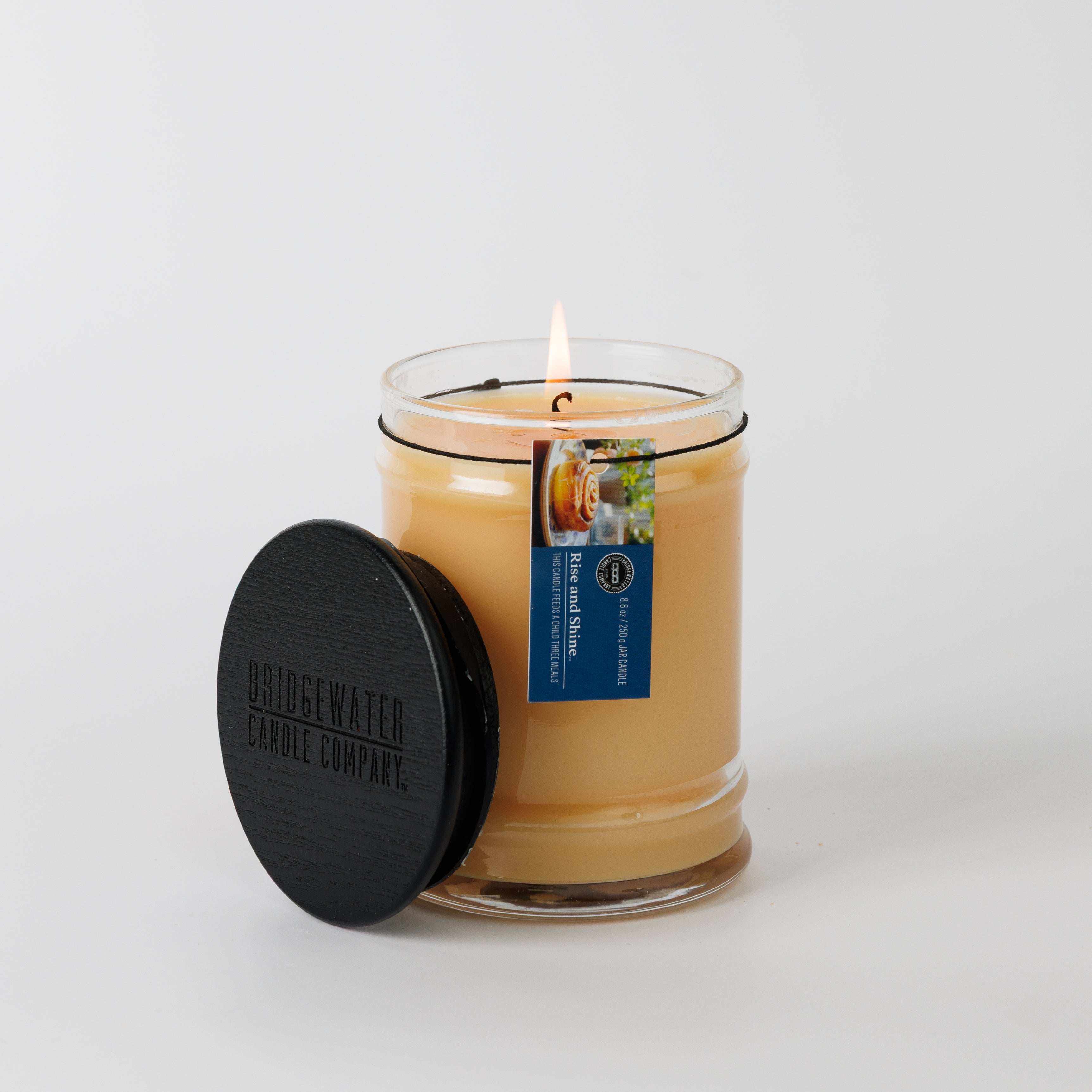 A lit Rise & Shine 8oz Jar Candle by Bridgewater Candle, with a black lid and blue tag, contains light beige soy-blend wax and signature fragrance. The candle sits against a plain white background.