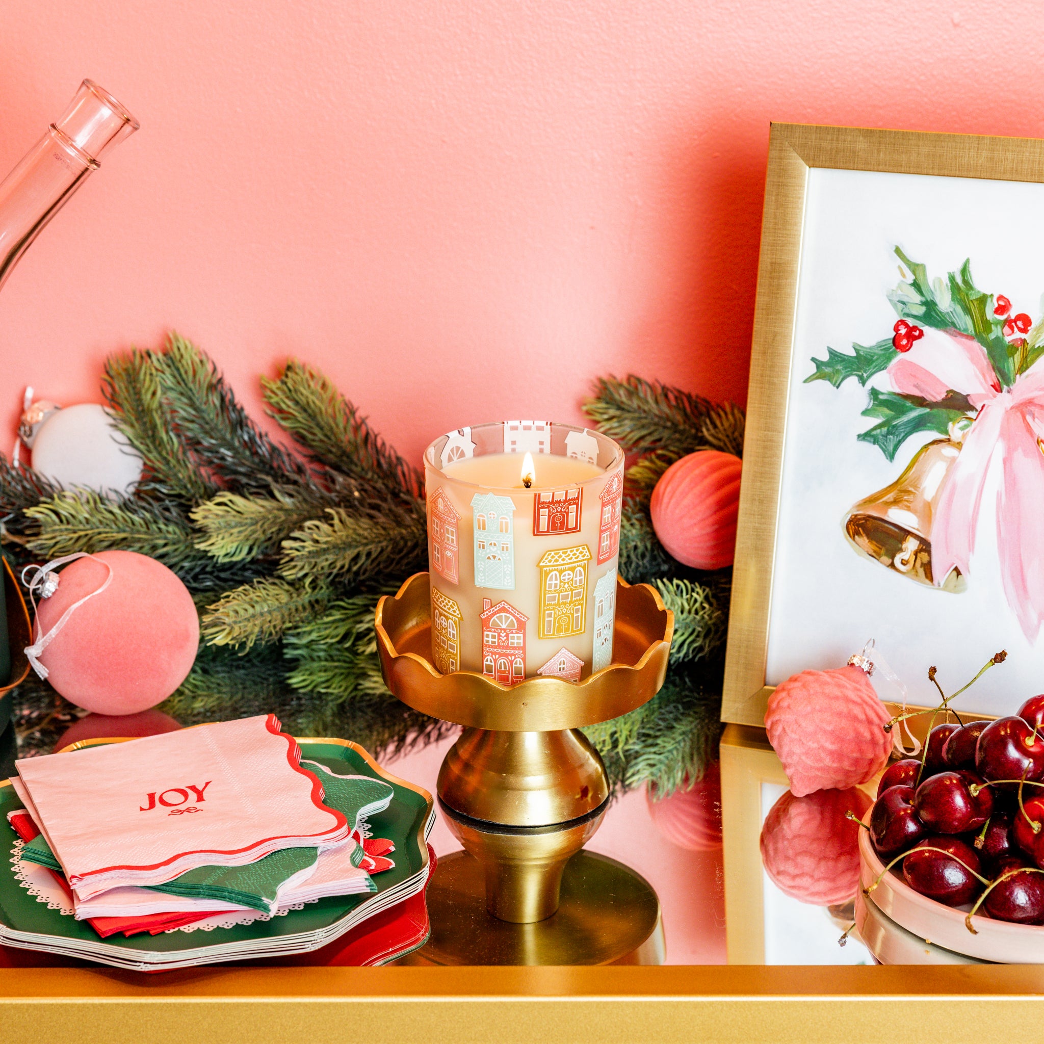 A festive display features the Bridgewater Candle Sweet Grace Holiday House Candle, evergreen branches, pink ornaments, a holly bell framed painting, decorative napkins, and cherries on a mirrored tray set against a pink wall.