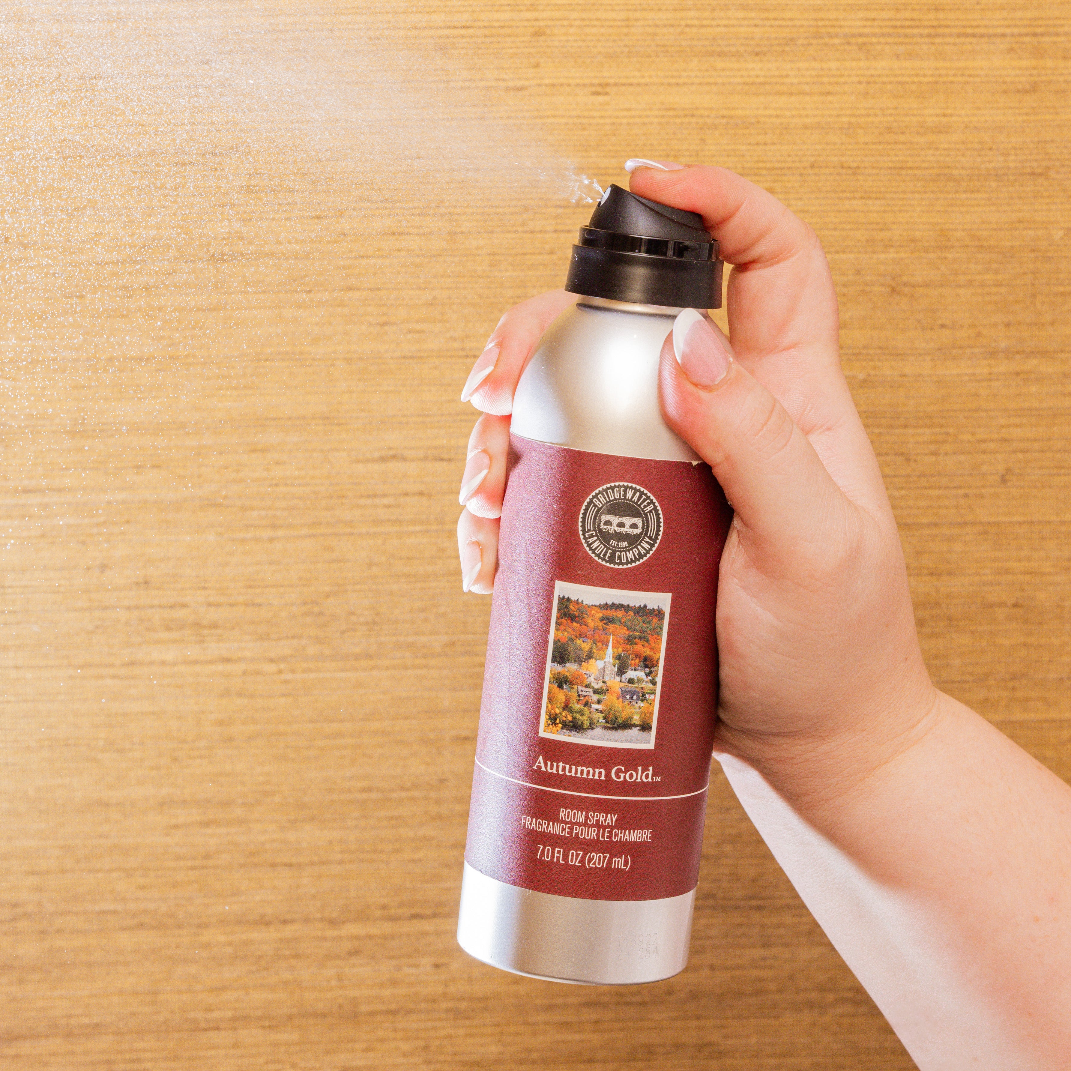 A hand sprays Bridgewater Candle’s Autumn Gold Room Spray against a wooden background, misting cozy fragrance from a 7 oz (207 ml) can adorned with an autumn landscape label.