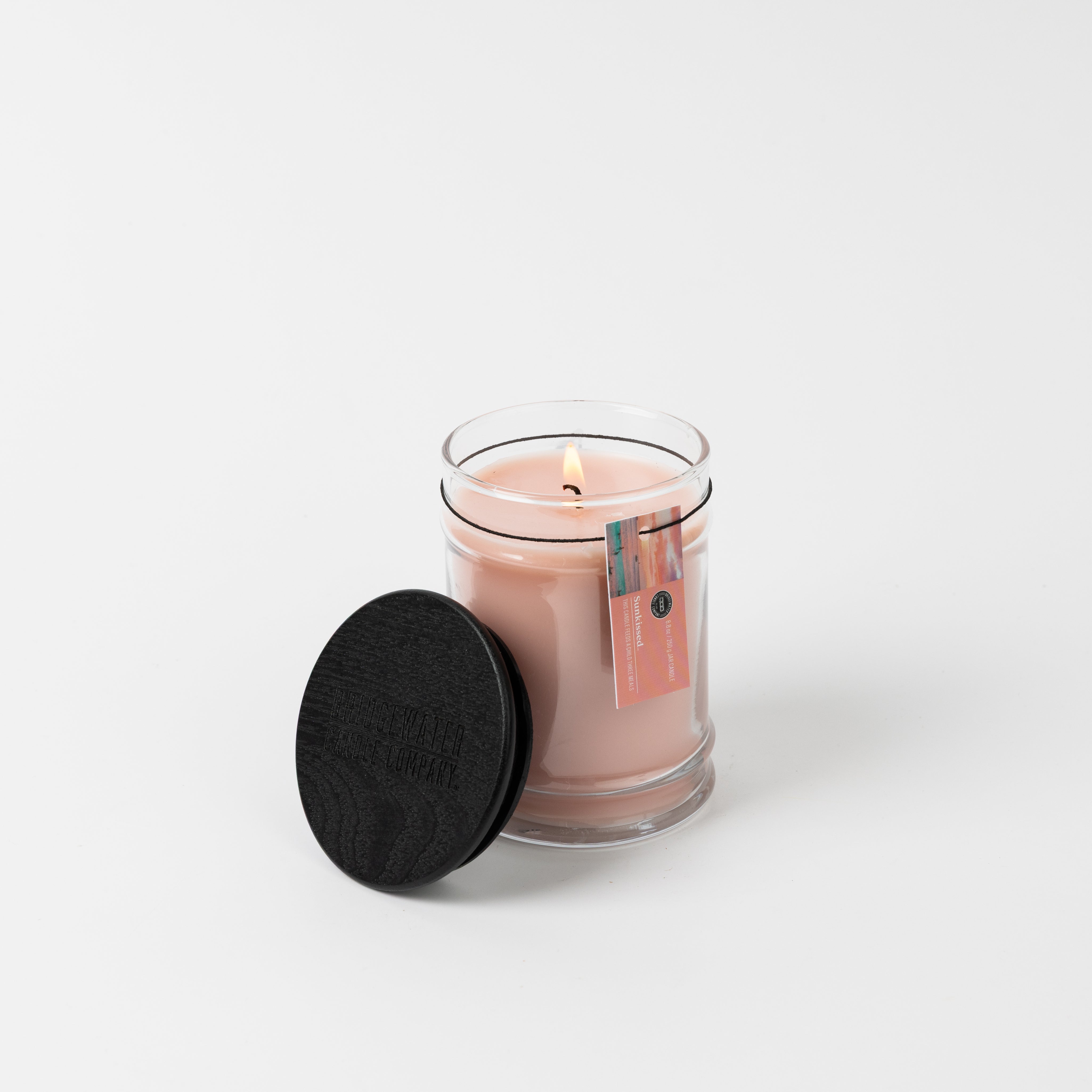 A lit Sunkissed 8oz Jar Candle by Bridgewater Candle, made with soy-blend wax, sits in a clear glass jar with a black lid beside it and a small tag attached. The background is white and minimalistic.