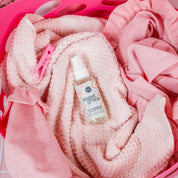 A bottle of Bridgewater Candle Sweet Grace Stain Remover - Travel Size rests on pink towels in a laundry basket with a pink clothespin nearby.