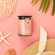The Sunkissed 8oz Jar Candle by Bridgewater Candle, made with soy-blend wax and a signature fragrance, rests in a glass jar with black lid on pink, surrounded by starfish, seashell, sunhat, green leaves, and netting for a beachy summer vibe.