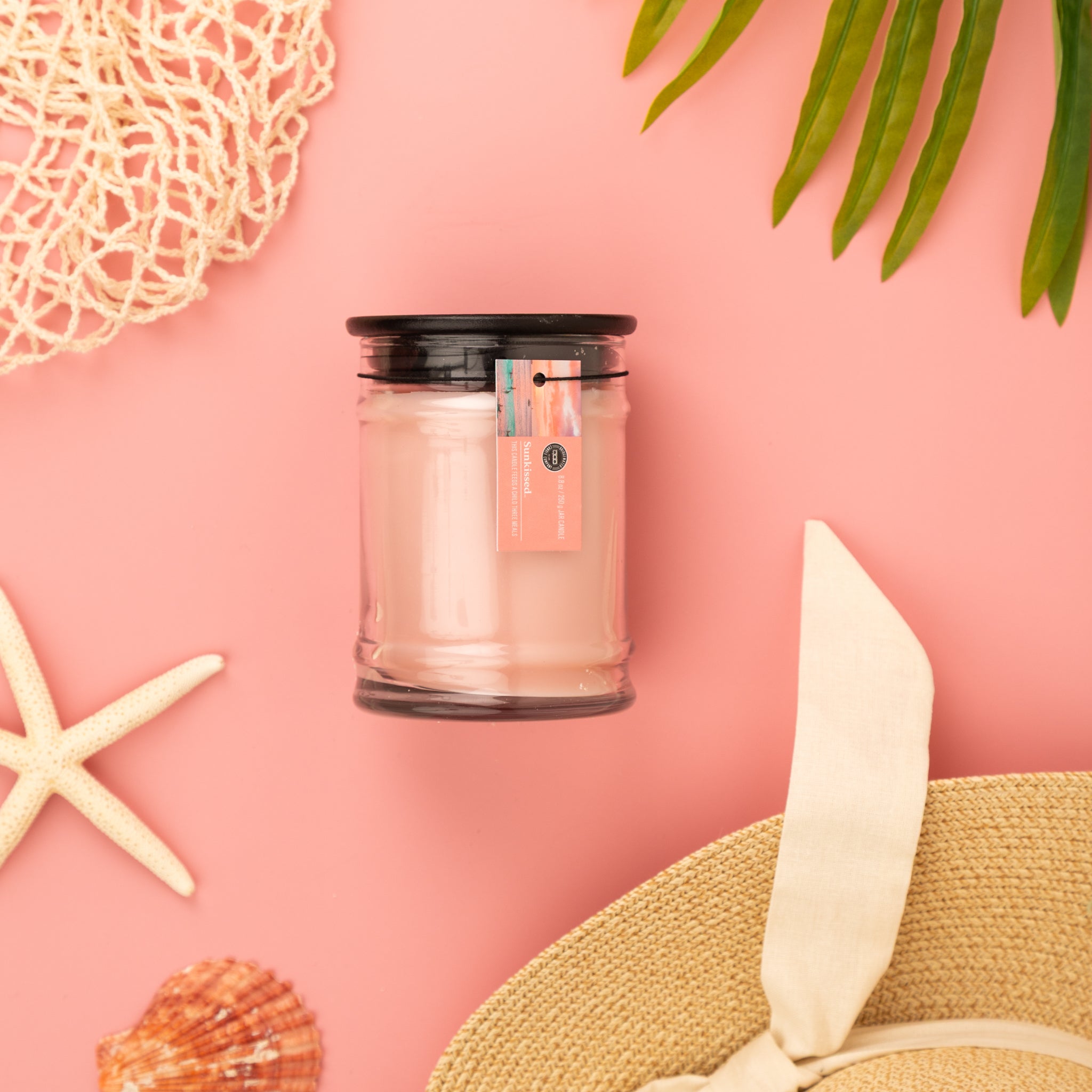 The Sunkissed 8oz Jar Candle by Bridgewater Candle, made with soy-blend wax and a signature fragrance, rests in a glass jar with black lid on pink, surrounded by starfish, seashell, sunhat, green leaves, and netting for a beachy summer vibe.
