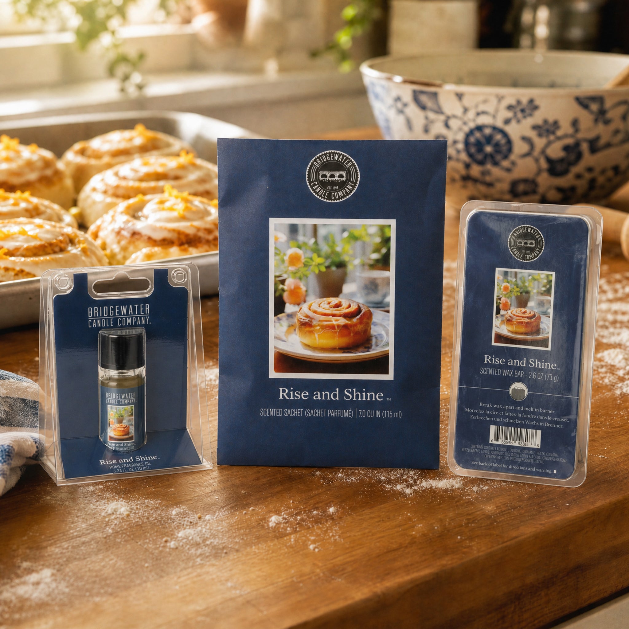 Bridgewater Candle’s Rise & Shine Flameless Bundle is shown on a floured kitchen counter with cinnamon rolls and a blue patterned mixing bowl in the background.
