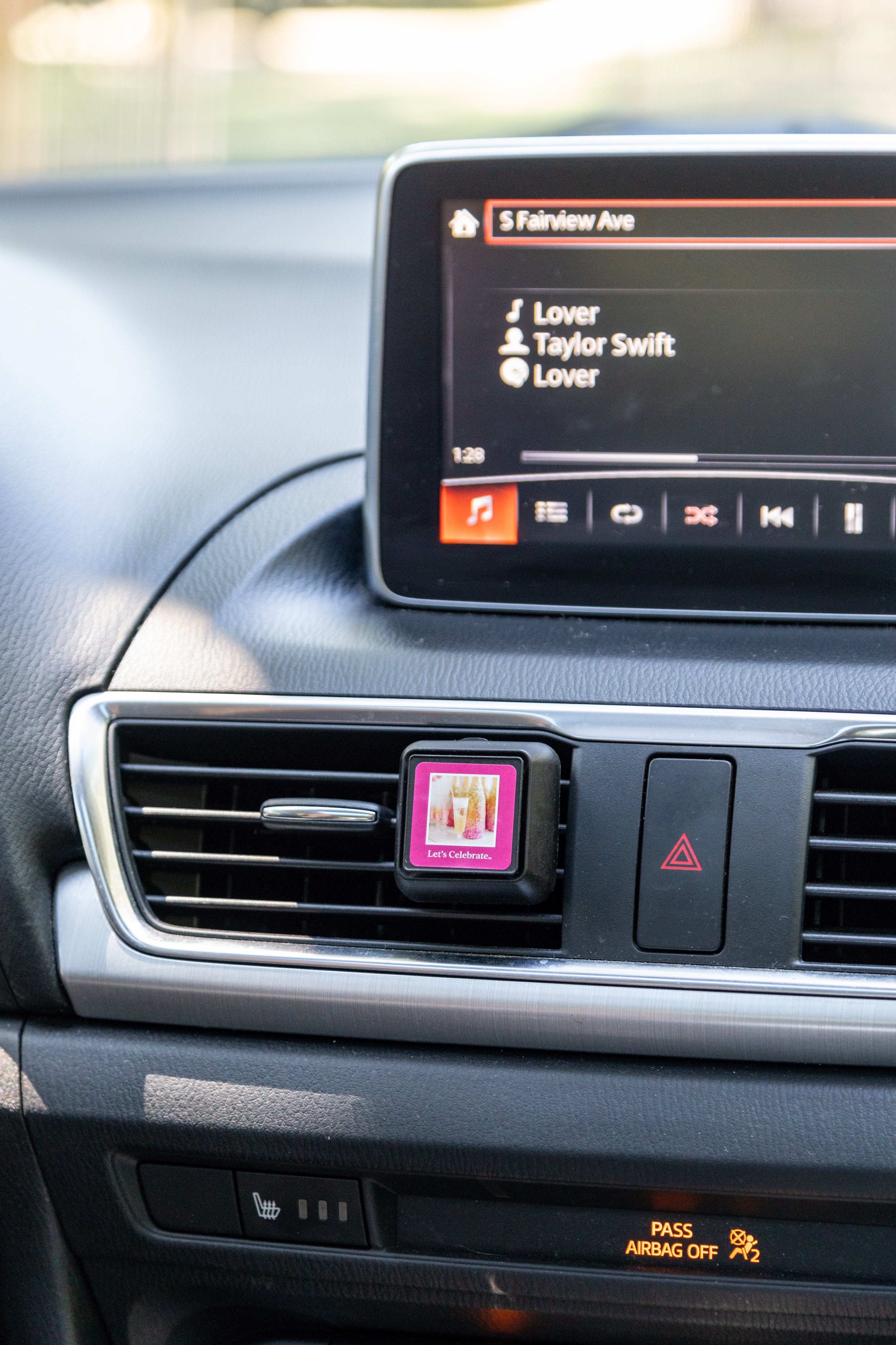 A square Auto Vent Clip-Let’s Celebrate by Bridgewater Candle attached to a dashboard, featuring a pink label with champagne glasses and the text Lets Celebrate.