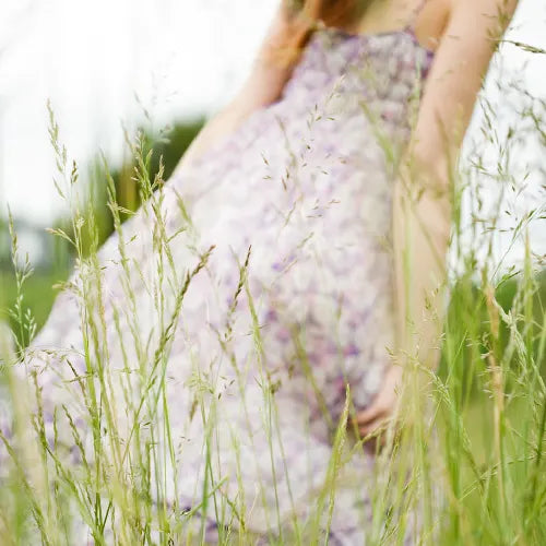 A person in a flowing, light purple floral dress stands in a grassy field, with tall green grass in the foreground and their face out of frame. The scene is soft-focused, creating a dreamy, peaceful atmosphere.