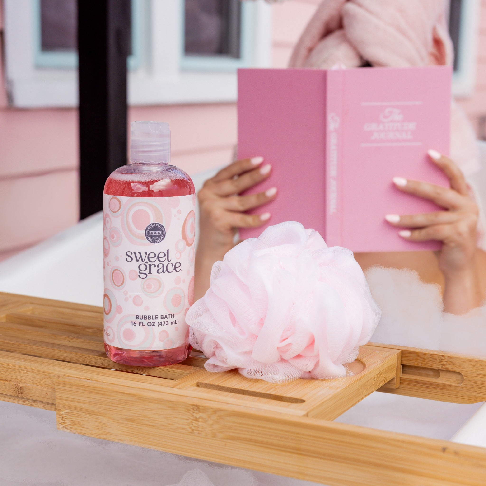 A bottle of Bridgewater Candle Sweet Grace Bubble Bath and a pink bath sponge rest on a wooden bathtub tray, with a person reading a book in the background.