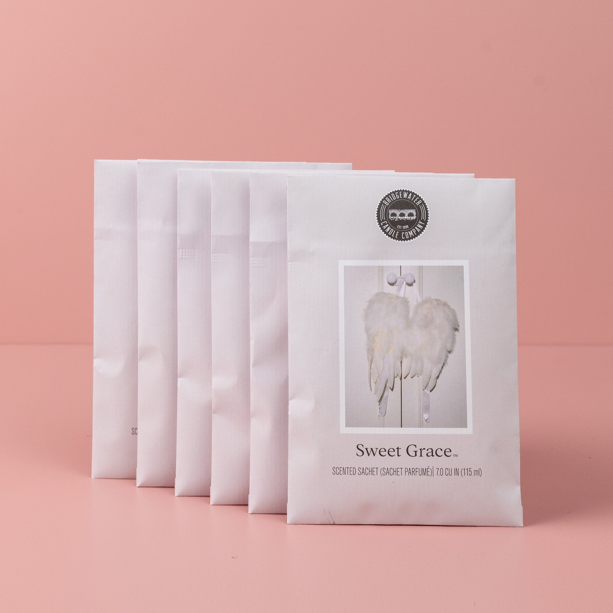 The Sweet Grace Sachet Bundle, with six sachets, our best-selling fragrance, stands upright in a row on a pink surface with a pink background. Each sachet displays angel wings and a round logo at the top.