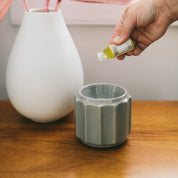 A hand holds a Fast Bundle Sweet Grace Home Fragrance Oil 3 Pack above a gray ceramic diffuser on a wooden table, offering a flameless scent experience. A white vase and pink leaves provide decorative charm in the background.