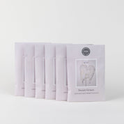 Six Sweet Grace Sachets are arranged in an arc on a white background. Each pastel pink sachet displays white angel wings, a circular top seal, and contains the brand’s best-selling fragrance.