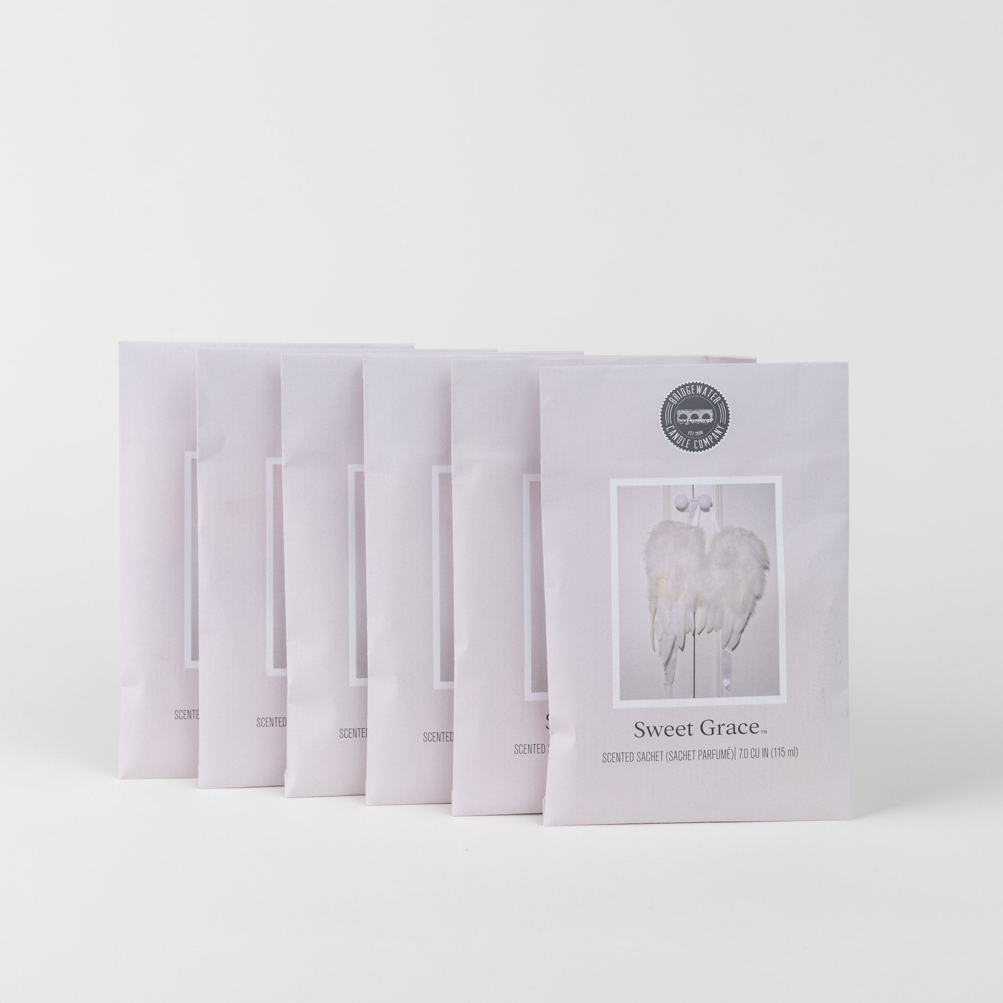 Six Sweet Grace Sachets are arranged in an arc on a white background. Each pastel pink sachet displays white angel wings, a circular top seal, and contains the brand’s best-selling fragrance.