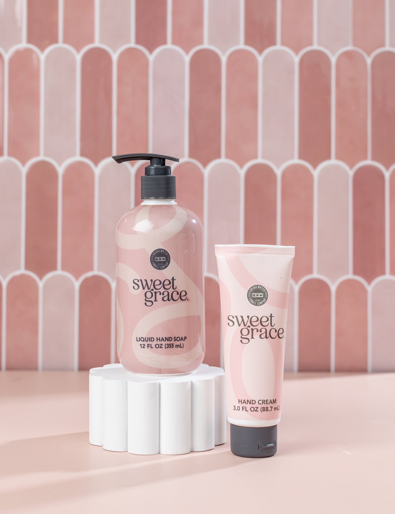 The Sweet Grace On Hand Bundle by Fast Bundle, featuring Hand Cream and Liquid Hand Soap, is displayed on a white pedestal with a pink tiled background—an ideal duo to elevate your handcare routine.