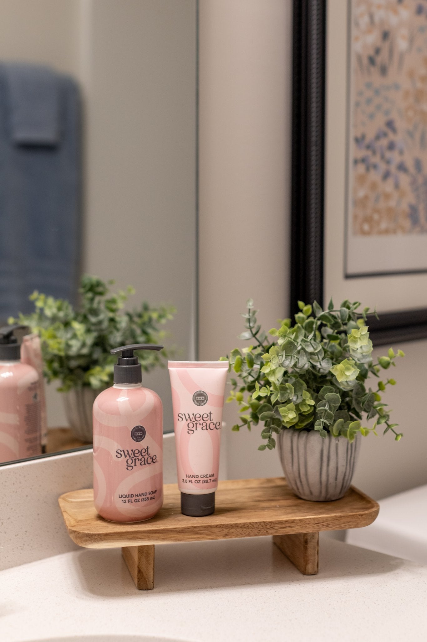 A wooden tray on the bathroom counter displays the Sweet Grace On Hand Bundle—Hand Cream & Liquid Hand Soap by Fast Bundle—plus a small potted green plant. A mirror and framed art complete this serene handcare setup.
