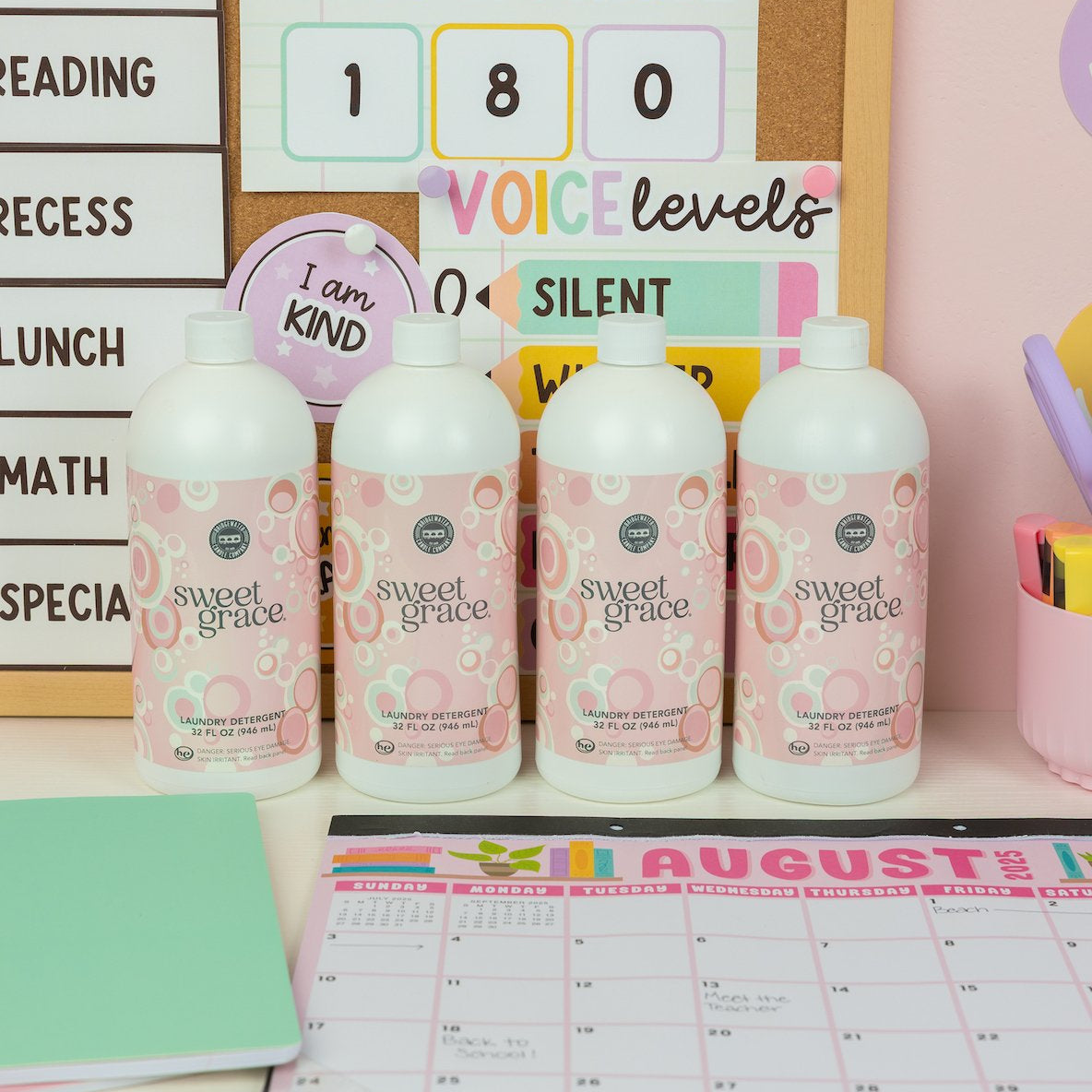 Four bottles from the Back to School Laundry Bundle 4pk by Fast Bundle are lined up on a desk alongside a calendar, notebooks, and classroom signs in the background, promising fresh-smelling laundry for every wash.