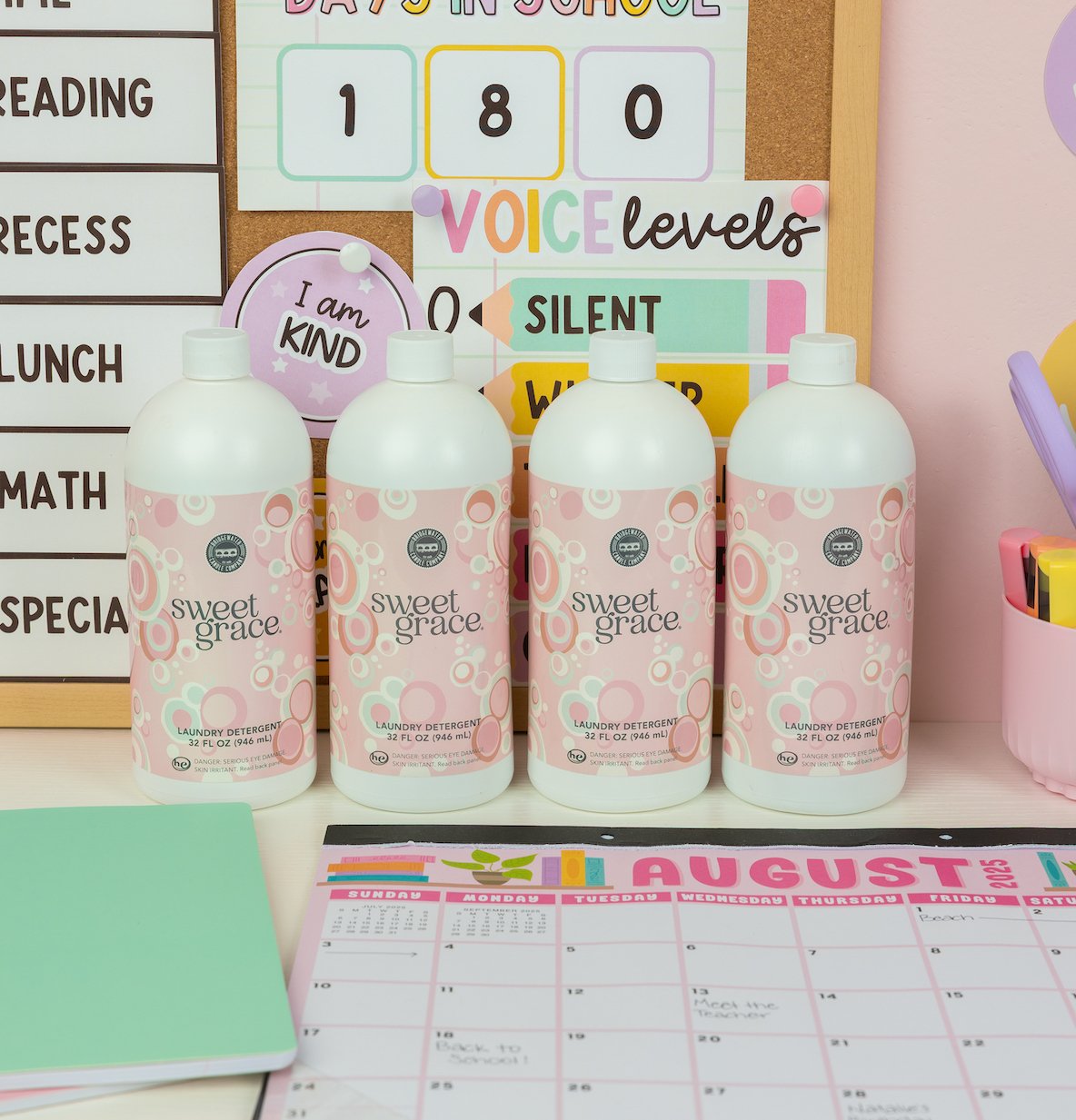 Four bottles from the Back to School Laundry Bundle 4pk by Fast Bundle are lined up on a desk alongside a calendar, notebooks, and classroom signs in the background, promising fresh-smelling laundry for every wash.
